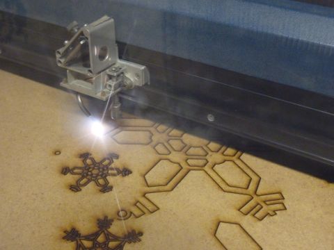 Laser cutting in progress. A sheet of MDF has partially cut snowflakes in it, with the laser head moving along the cut line and cutting further.
